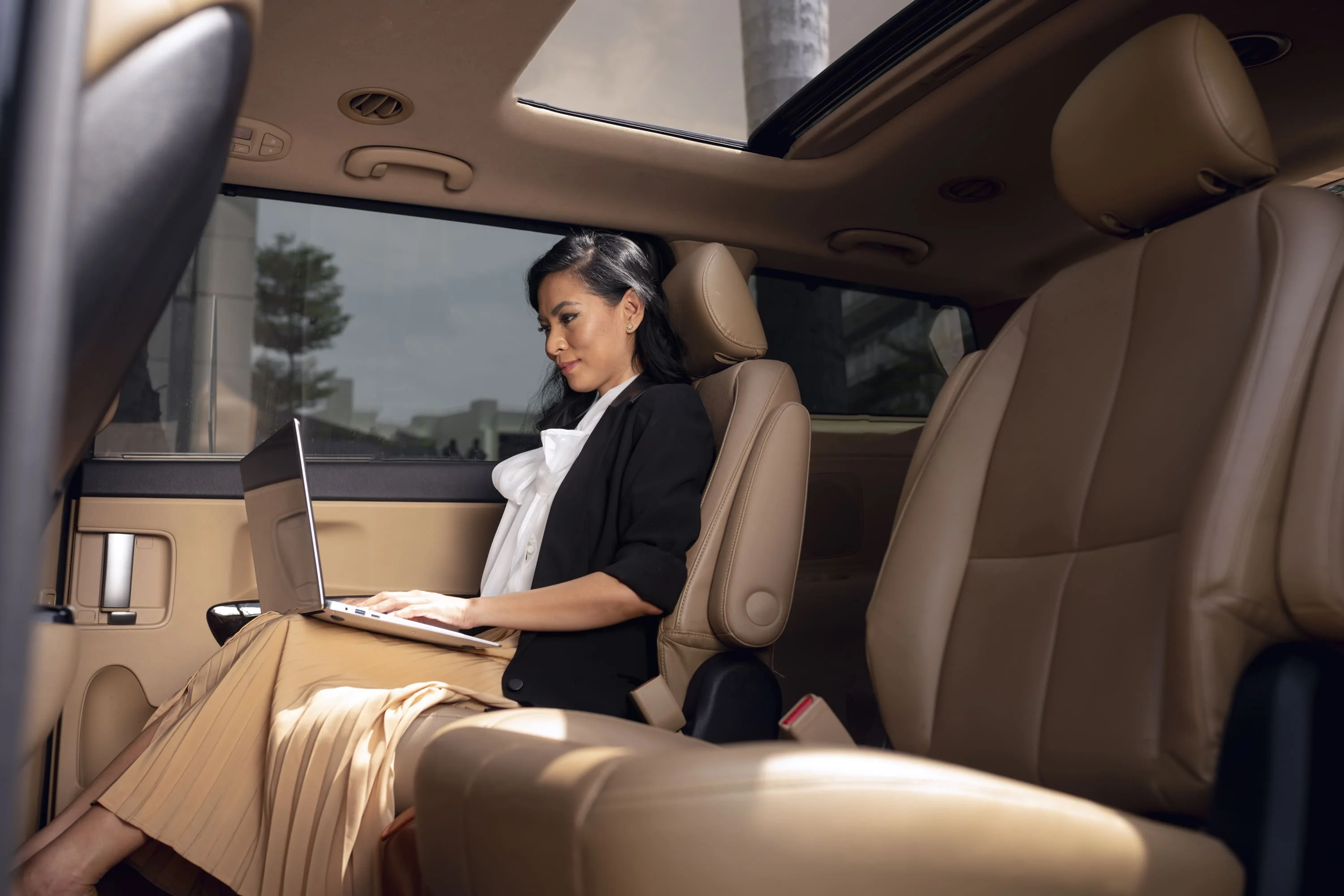 A passenger working on a laptop in the cream leather rear seat of a chauffeured vehicle